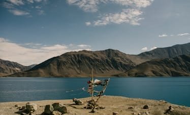 A tranquil mountain lake with Tibetan prayer flags waving in the wind, under a bright blue sky.