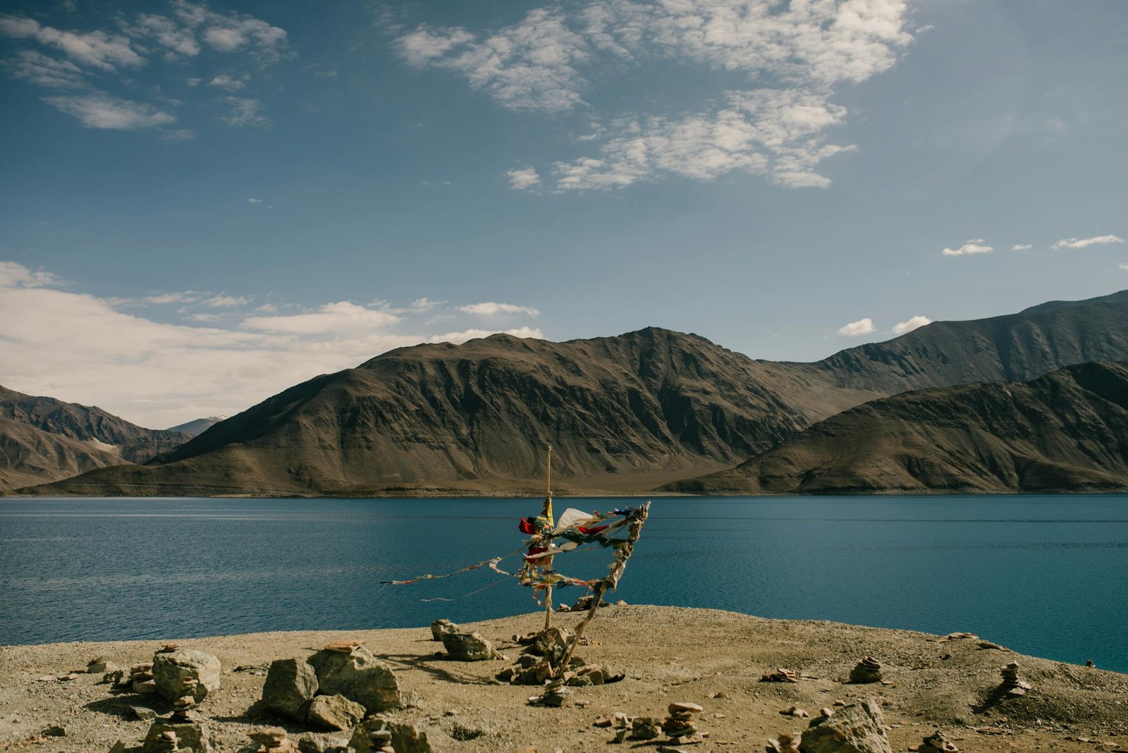 A tranquil mountain lake with Tibetan prayer flags waving in the wind, under a bright blue sky.
