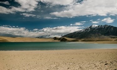 Sandy beach near river against snowy mountain peak and blue sly with clouds in nature on sunny day in countryside