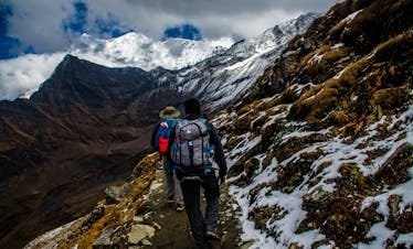 Two hikers navigating a snowy trail in rugged mountainous terrain under dramatic skies.