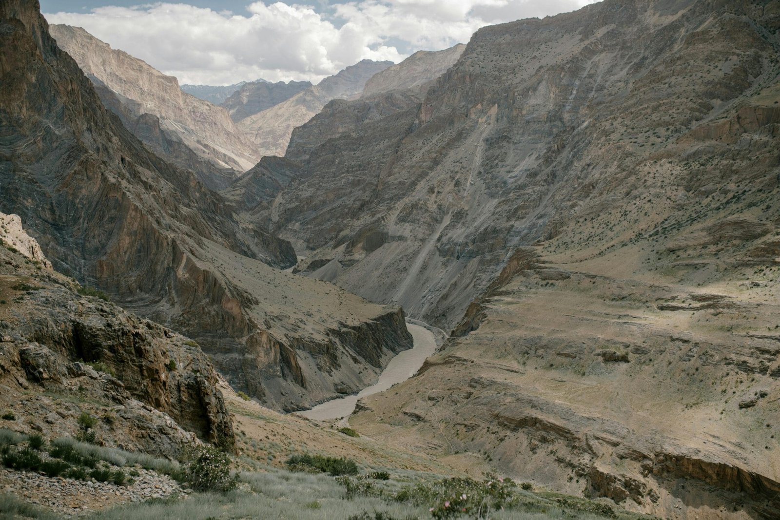 Picturesque landscape of narrow river flowing among rough rocky mountain range covered with dry vegetation and sand against cloudy sky