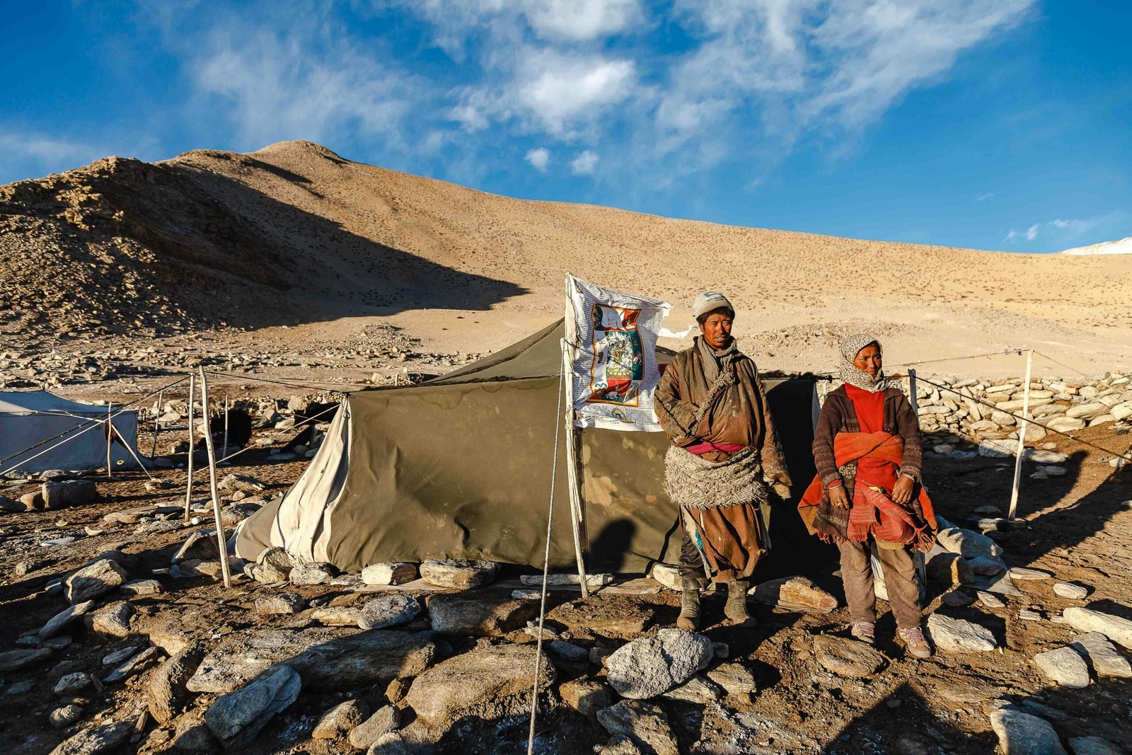 Two nomadic individuals stand by a tent in Ladakh, India's stark desert landscape.